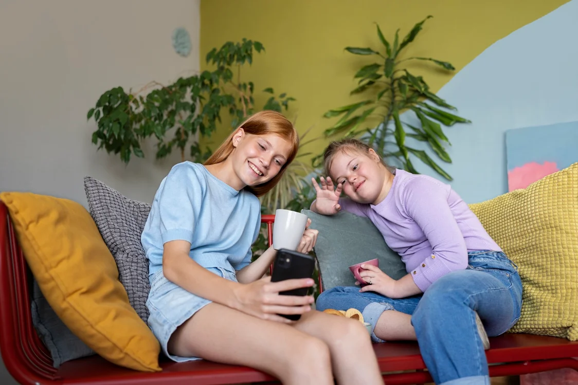 Two young women relaxing together on a sofa, smiling at a phone.