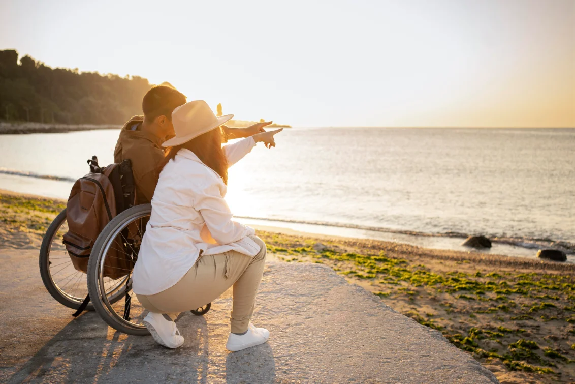 A support coordinator and a participant using a wheelchair, pointing out at the sea together.